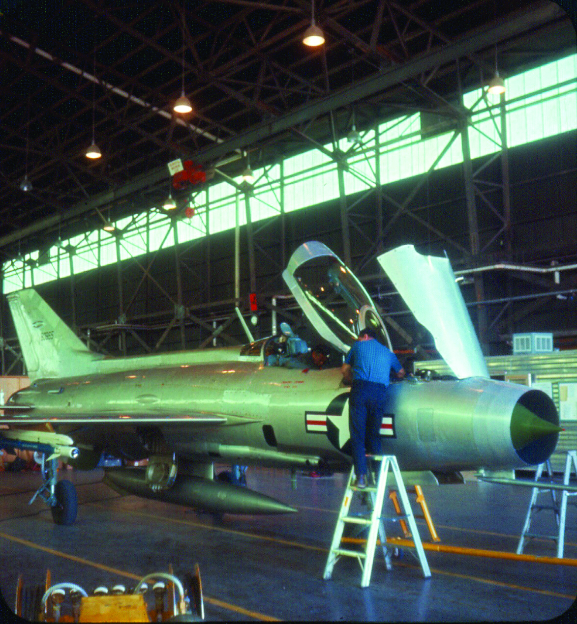 Technicians work on a silver MiG-21 fighter jet inside a large aircraft hangar. One technician stands on a ladder near the open cockpit. The aircraft displays a U.S. insignia.